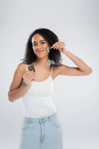 Erfreut afrikanisch-amerikanische Frau hält Gesichtsschaber und verwendet Jade-Rolle isoliert auf grau — Stockfoto