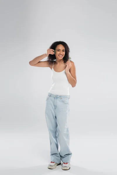 Longitud completa de la sonriente mujer afroamericana en camiseta sin mangas y jeans posando con exfoliante facial y rodillo de jade sobre fondo gris - foto de stock