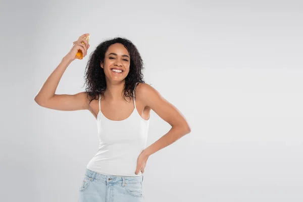 Усміхнена афроамериканська жінка наносить масло для волосся, стоячи з рукою на стегно ізольовано на сірому — стокове фото