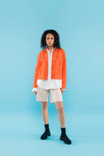 Volle Länge der afrikanisch-amerikanischen Frau in stilvoller orangefarbener Jacke und Baumwoll-Shorts, die in die Kamera auf blauem Hintergrund schaut — Stockfoto