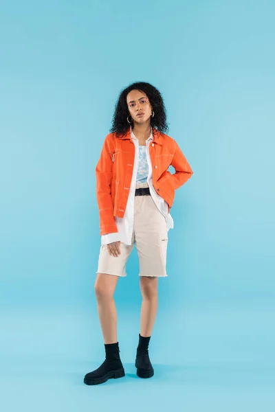 Повна довжина афроамериканської жінки в яскраво-помаранчевій куртці і бавовняних шортах позує з рукою на стегні на синьому фоні — стокове фото