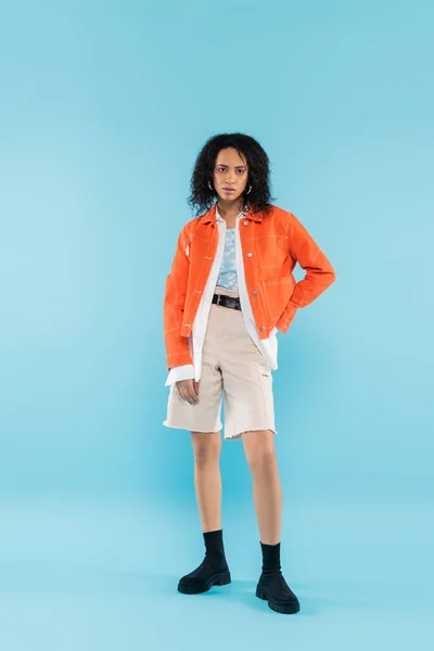 Повна довжина афроамериканської жінки в бавовняних шортах і помаранчевій куртці позує з рукою на стегні на синьому фоні — стокове фото