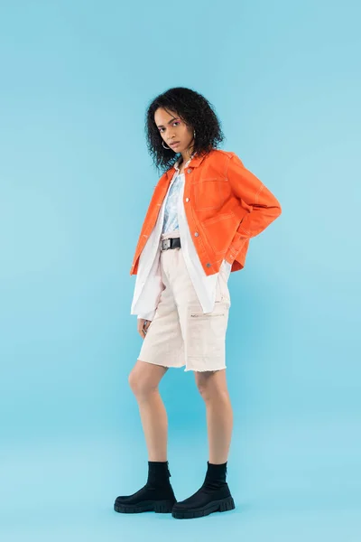 Longitud completa de la mujer afroamericana de moda en chaqueta naranja posando con la mano en el bolsillo trasero de pantalones cortos sobre fondo azul - foto de stock