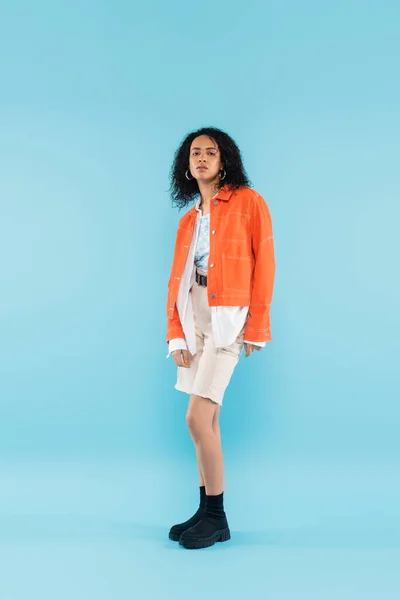 Longitud completa de la mujer afroamericana en chaqueta naranja y pantalones cortos blancos de pie sobre fondo azul - foto de stock