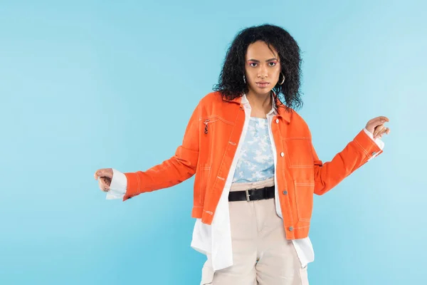 Brunette african american woman in bright orange jacket looking at camera isolated on blue — Stock Photo