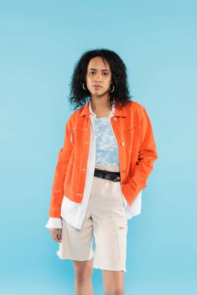 Brunette african american woman in bright orange jacket posing with hand in pocket of cotton shorts isolated on blue — Stock Photo