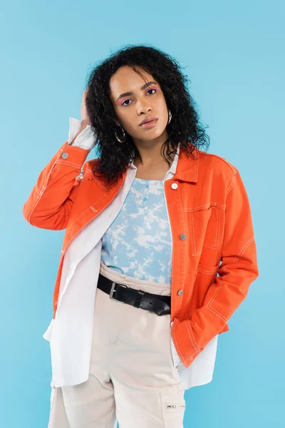 Brunette african american woman in trendy jacket posing with hand in pocket of cotton shorts isolated on blue — Stock Photo