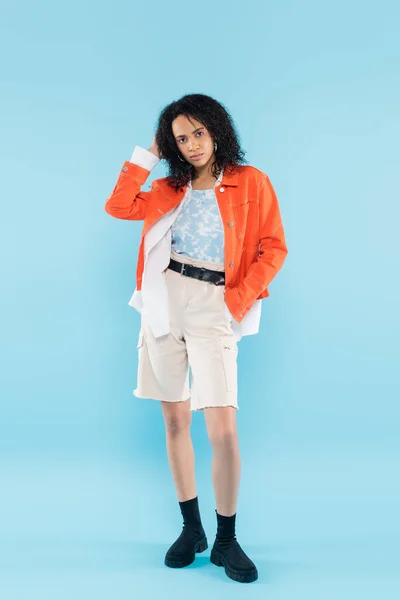 Full length of stylish african american woman standing with hand in pocket of cotton shorts and touching wavy hair on blue background — Stock Photo