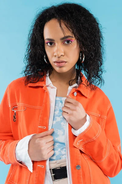 Porträt einer brünetten Afroamerikanerin in leuchtend orangefarbener Jacke, die isoliert auf blauem Grund in die Kamera blickt — Stockfoto