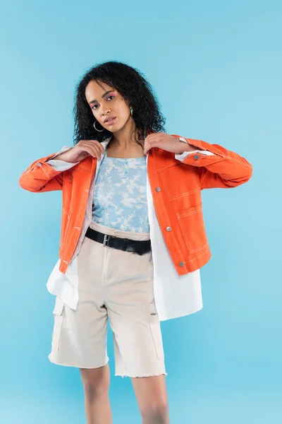 Curly african american woman in white shorts adjusting bright orange jacket isolated on blue — Stock Photo