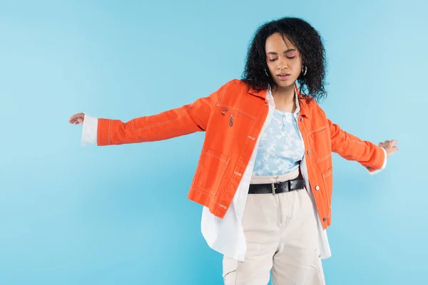 Trendy african american woman in orange jacket and shorts posing with outstretched hands isolated on blue — Stock Photo
