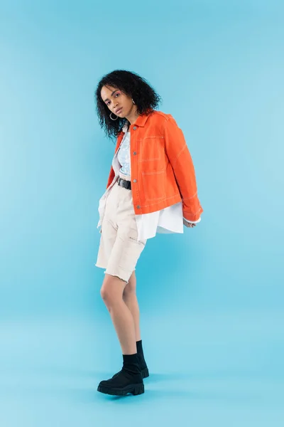 Volle Länge der brünetten afrikanisch-amerikanische Frau in orangefarbener Jacke und schwarzen Stiefeln Blick in die Kamera auf blauem Hintergrund — Stockfoto