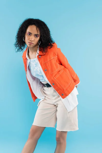 African american woman with curly brunette hair posing in orange jacket and cotton shorts isolated on blue — Stock Photo