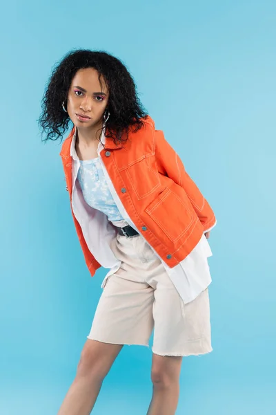 Mulher americana africana com cabelo morena encaracolado posando em jaqueta laranja e shorts de algodão isolados em azul — Fotografia de Stock