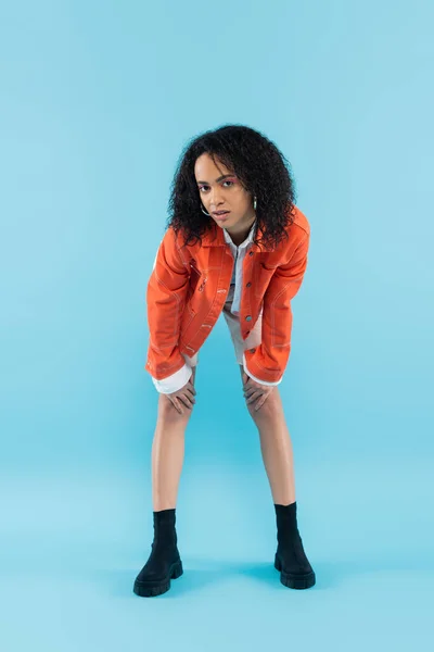 Повна довжина кучерявої афроамериканської жінки в модній помаранчевій куртці позує і дивиться на камеру на синьому фоні — стокове фото