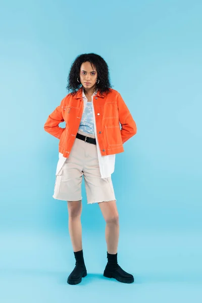 Piena lunghezza della donna afroamericana alla moda in pantaloncini bianchi e giacca arancione in posa con le mani sui fianchi su sfondo blu — Stock Photo