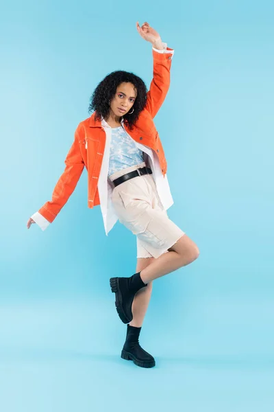 Piena lunghezza della donna afroamericana alla moda in giacca arancione brillante e stivali neri in posa con mano sollevata su sfondo blu — Foto stock