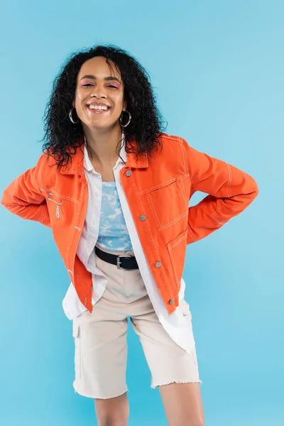 Cheerful african american woman in stylish orange jacket smiling at camera isolated on blue — Stock Photo