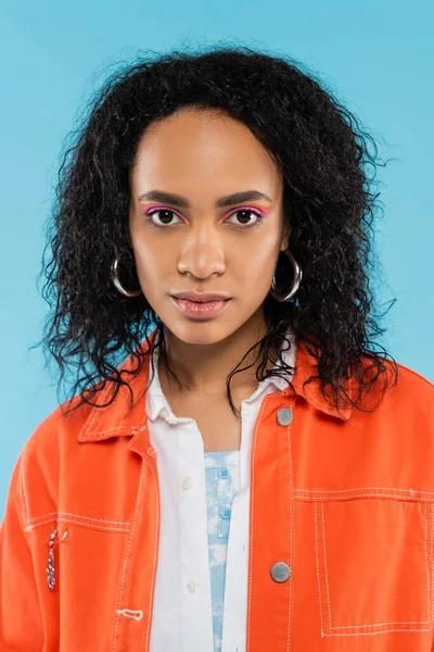 Retrato de mujer afroamericana con cabello ondulado y maquillaje brillante mirando a la cámara aislada en azul - foto de stock