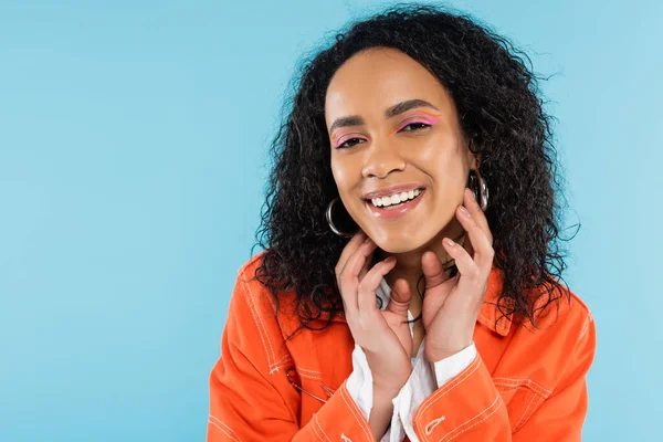 Porträt einer glücklichen afrikanisch-amerikanischen Frau mit hellem Make-up, die Hände in Gesichtsnähe hält, isoliert auf blauem Grund — Stockfoto