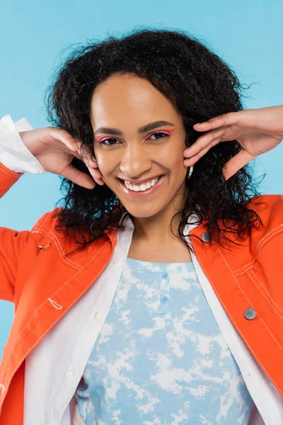 Retrato de alegre y elegante mujer afroamericana con maquillaje brillante cogido de la mano cerca de la cara aislado en azul - foto de stock