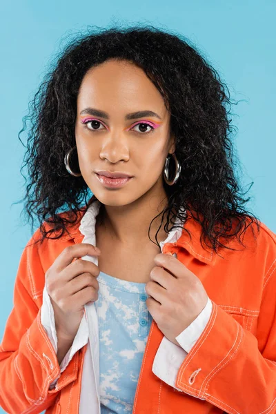Porträt einer brünetten afrikanisch-amerikanischen Frau mit hellem Make-up und Ohrringen, die isoliert auf blauem Grund in die Kamera blickt — Stockfoto