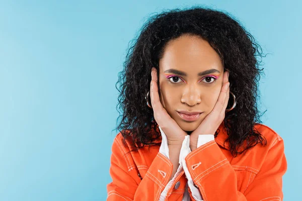Porträt einer brünetten afrikanisch-amerikanischen Frau in orangefarbener Jacke, die das Gesicht berührt und isoliert auf blau in die Kamera blickt — Stockfoto