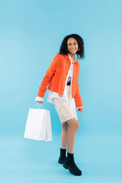 Повна довжина задоволеної афроамериканської жінки в помаранчевій куртці і чоботях, що тримають білі сумки для покупок на синьому фоні — стокове фото