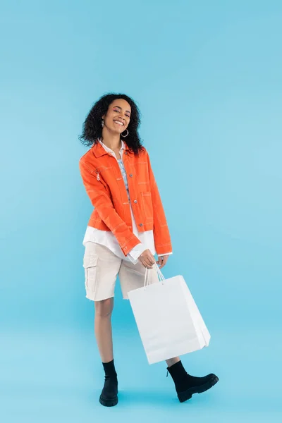 Comprimento total de mulher americana africana alegre em jaqueta laranja e shorts de algodão posando com sacos de compras no fundo azul — Fotografia de Stock
