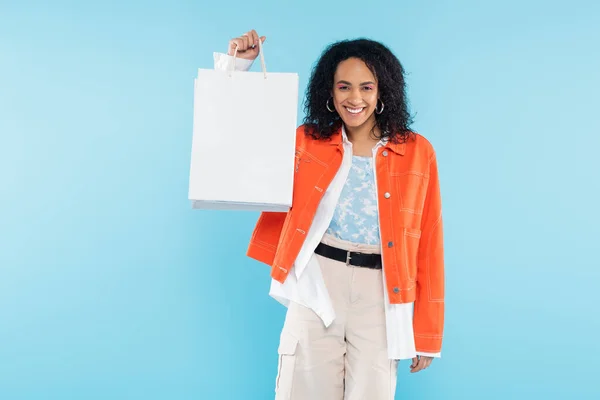 Overjoyed african american woman in orange jacket showing white shopping bags isolated on blue — Stock Photo