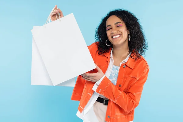 Super alegre mulher americana africana em jaqueta laranja segurando sacos de compras brancos isolados em azul — Fotografia de Stock