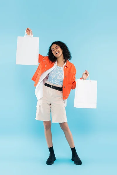 Comprimento total de mulher americana africana alegre e na moda posando com sacos de compras brancos no fundo azul — Fotografia de Stock