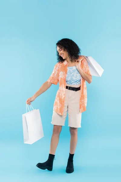 Longitud completa de la mujer afroamericana de moda en pantalones cortos y botas negras de pie con bolsas de compras sobre fondo azul - foto de stock