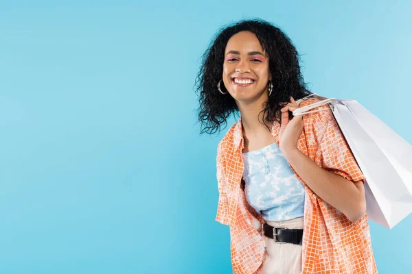 Mujer afroamericana elegante con el pelo moreno rizado sosteniendo bolsas de compras y sonriendo a la cámara aislada en azul - foto de stock