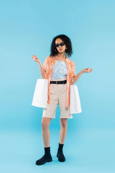 Pleine longueur de femme afro-américaine en lunettes de soleil et short posant avec des sacs à provisions blancs sur fond bleu — Photo de stock