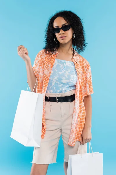 Mujer afroamericana de moda en gafas de sol posando con bolsas de compras aisladas en azul - foto de stock