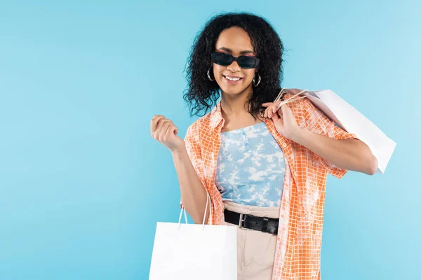 Mujer afroamericana de moda en gafas de sol de pie con bolsas de compras blancas aisladas en azul - foto de stock