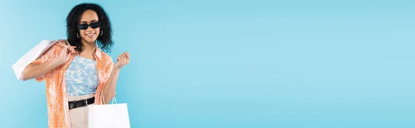 Donna afroamericana sorridente in abiti alla moda e occhiali da sole che tiene borse della spesa bianche isolate su blu, banner — Foto stock