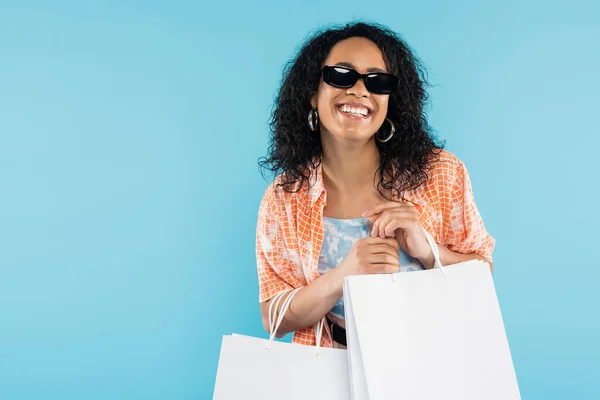 Donna afro-americana felicissima con eleganti occhiali da sole che tengono borse bianche isolate sul blu — Foto stock