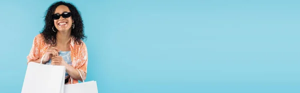 Улыбающаяся африканская американка в модной одежде и солнцезащитных очках позирует с мешками для покупок изолированными на голубом, баннер — стоковое фото