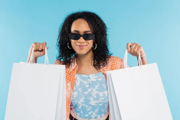Femme afro-américaine à la mode dans des lunettes de soleil montrant des sacs à provisions blancs isolés sur bleu — Photo de stock