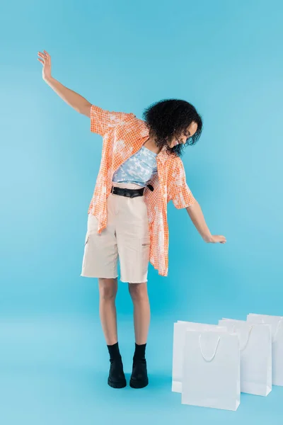 Piena lunghezza di felice donna africana americana in stivali neri e pantaloncini guardando borse della spesa bianche su sfondo blu — Foto stock