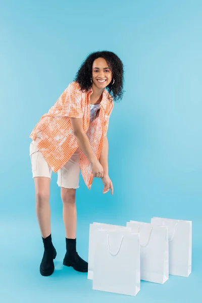 Longitud completa de la mujer afroamericana complacida mirando a la cámara y apuntando a las bolsas de compras blancas sobre fondo azul - foto de stock