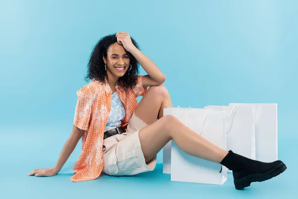 Longitud completa de la elegante mujer afroamericana sentada cerca de bolsas de compras y sonriendo a la cámara sobre fondo azul - foto de stock