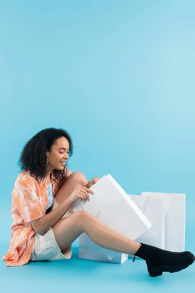 Volle Länge der zufriedenen afrikanisch-amerikanischen Frau, die in Einkaufstasche schaut, während sie auf blauem Hintergrund sitzt — Stockfoto