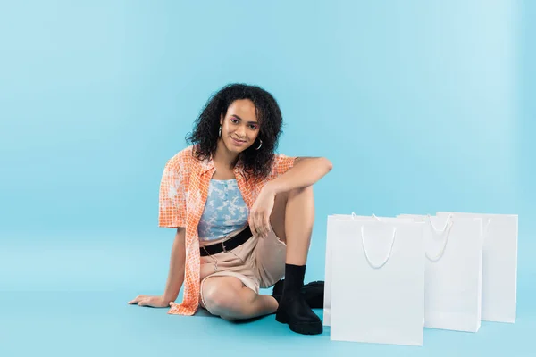 Longitud completa de la sonriente mujer afroamericana en ropa de moda sentado cerca de bolsas de compras sobre fondo azul — Stock Photo