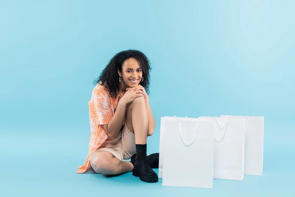 Comprimento total de mulher americana africana sorridente em roupas da moda sentado perto de sacos de compras brancos no fundo azul — Fotografia de Stock
