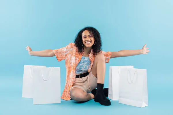 Volle Länge der fröhlichen afrikanisch-amerikanischen Frau sitzt mit ausgestreckten Händen neben weißen Einkaufstaschen auf blauem Hintergrund — Stockfoto