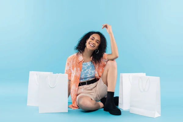 Longitud completa de morena mujer afroamericana sonriendo a la cámara mientras está sentado cerca de bolsas de compras sobre fondo azul - foto de stock