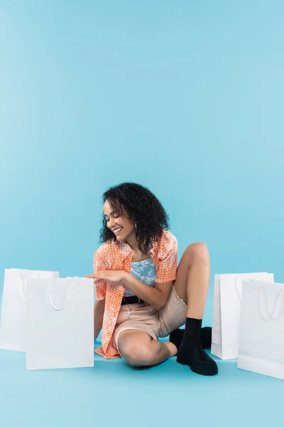 Volle Länge der fröhlichen und trendigen afrikanisch-amerikanischen Frau sitzt neben weißen Einkaufstaschen auf blauem Hintergrund — Stockfoto